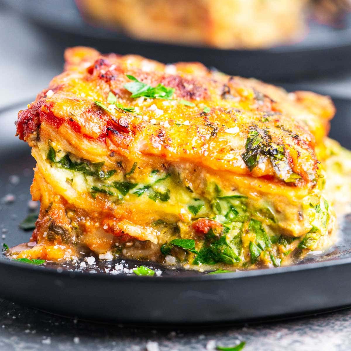 A close-up of a slice of spinach and ricotta lasagna, featuring layers of spinach, melted cheese, tomato sauce, and herbs, served on a dark plate.