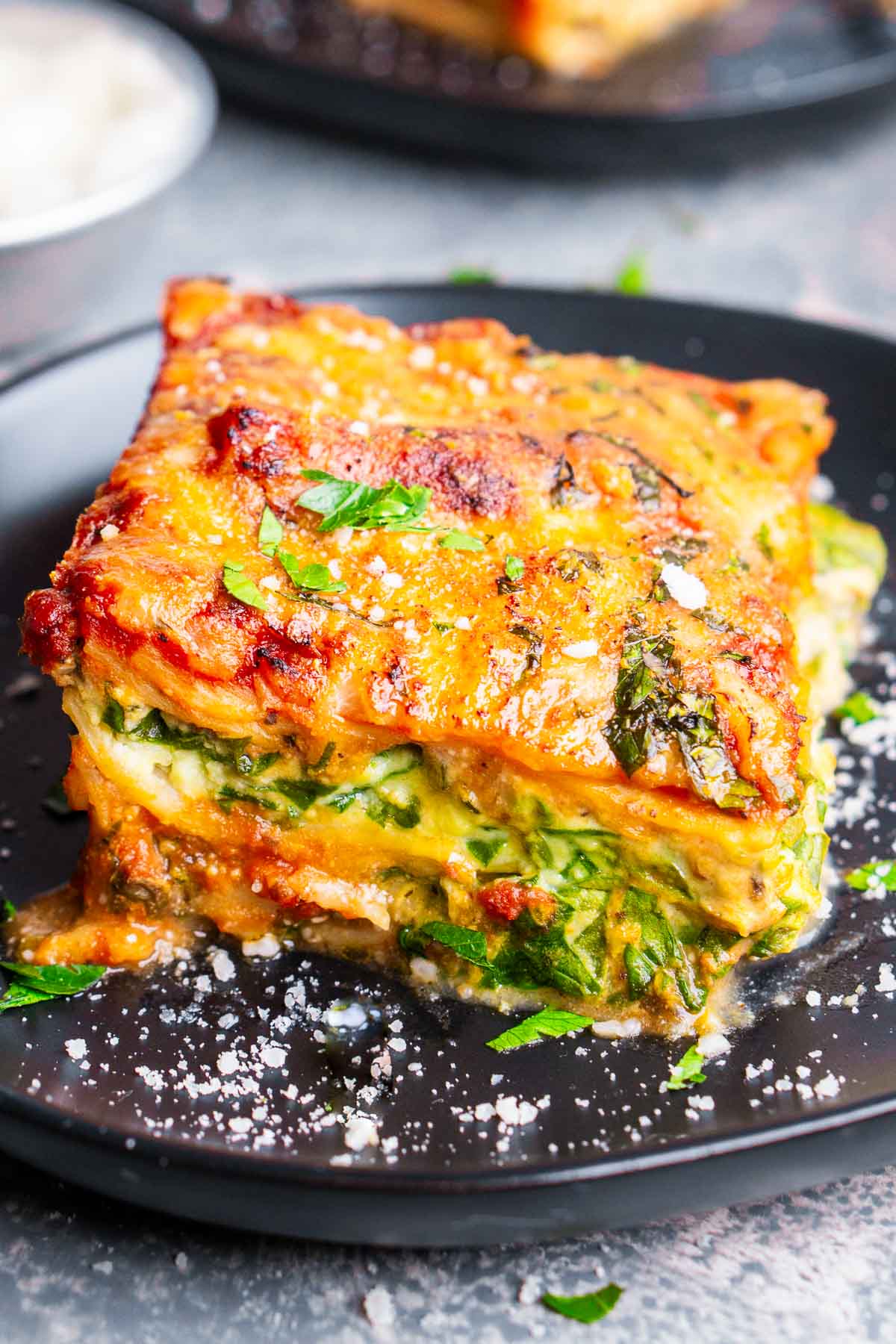 A close-up of a slice of spinach and ricotta lasagna, featuring layers of spinach, melted cheese, tomato sauce, and herbs, served on a dark plate.