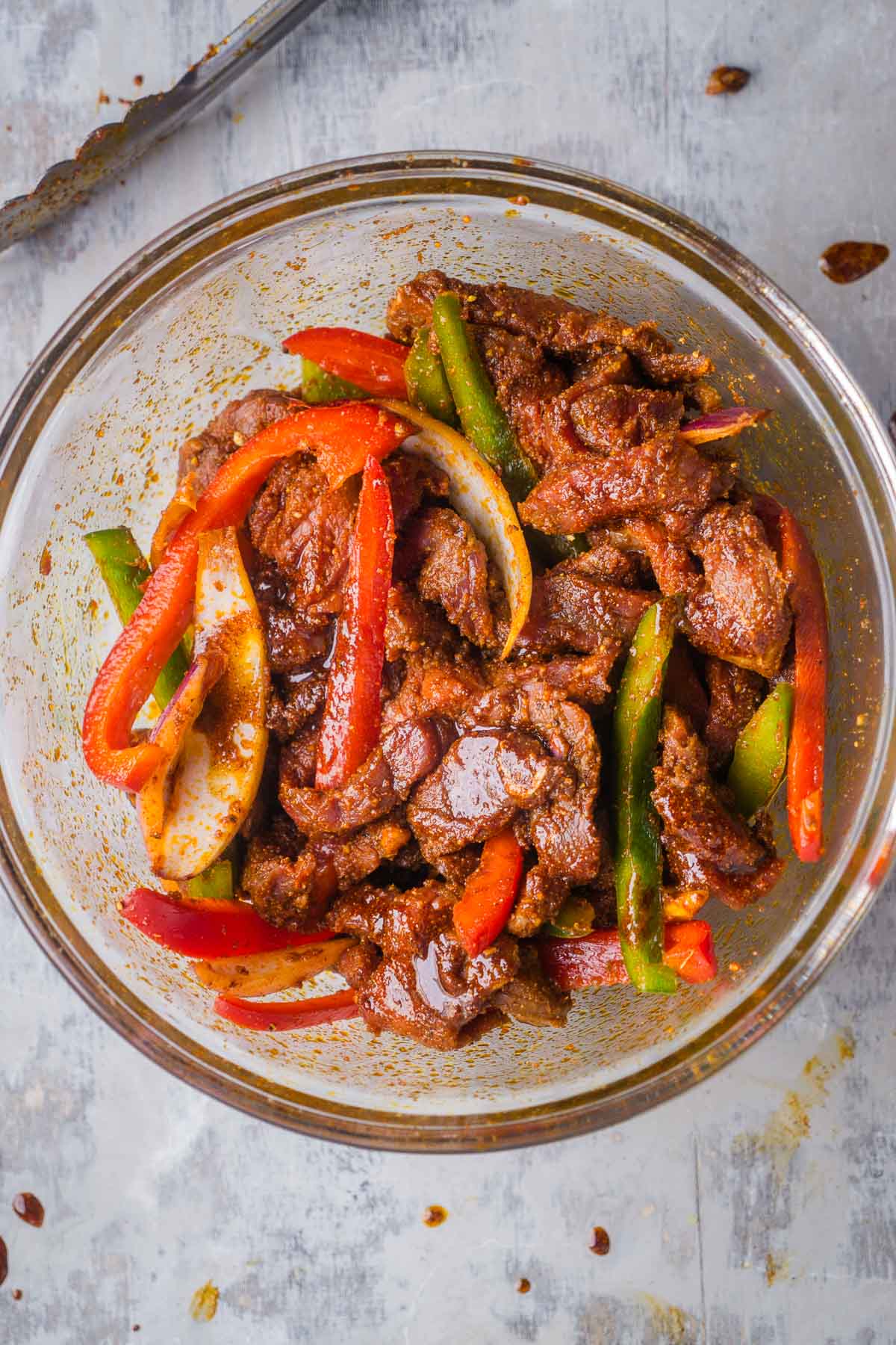 Sliced steak, bell peppers, and onions coated with fajita marinade in glass mixing bowl.