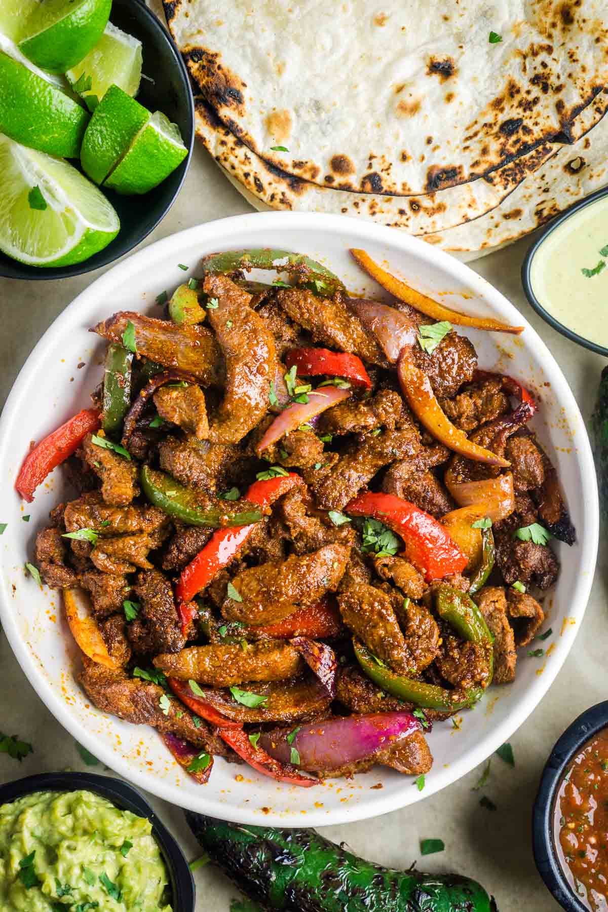 Juicy cooked steak fajitas in white bowl next to charred tortillas, lime wedges, guacamole, salsa, and charred jalapeno.