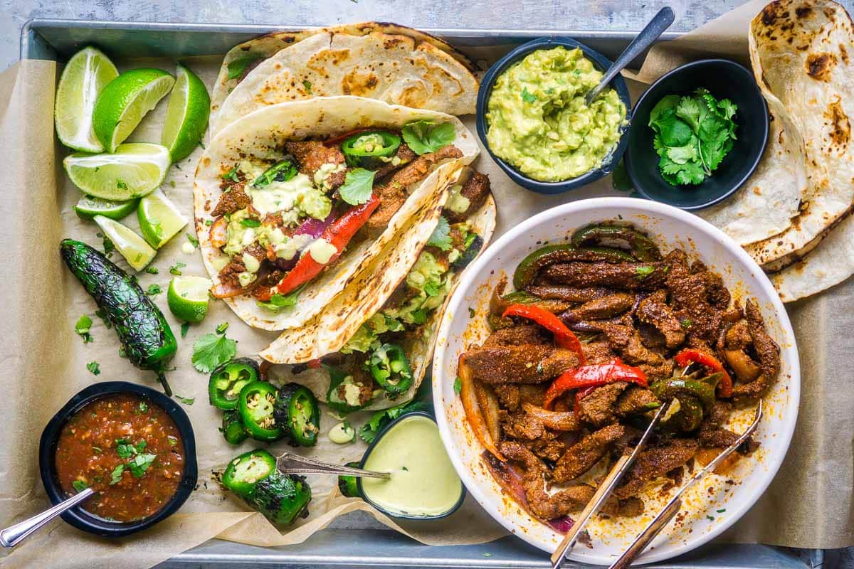 Steak fajitas in flour tortillas on tray next to bowls of Mexican toppings.