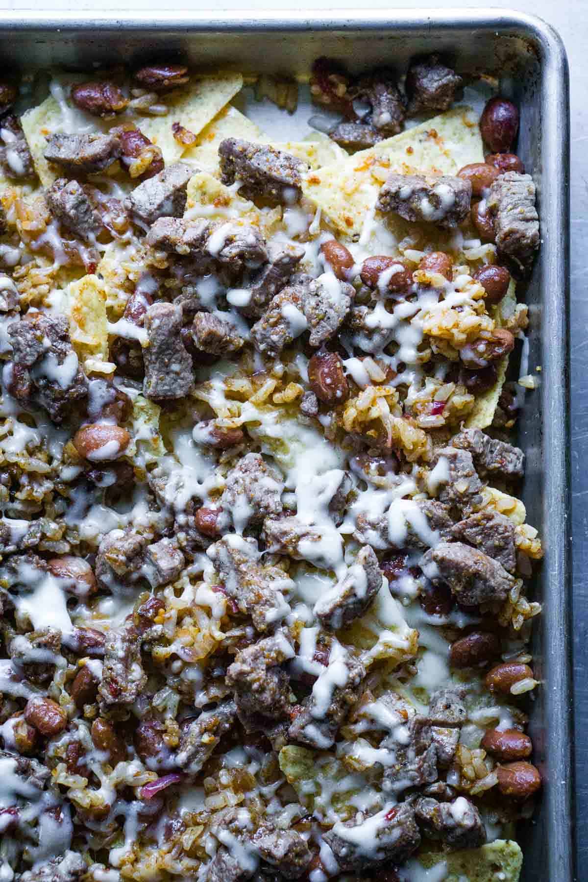 tortilla chips on baking sheet with whole pinto beans, rice, steak bites, and melted cheese