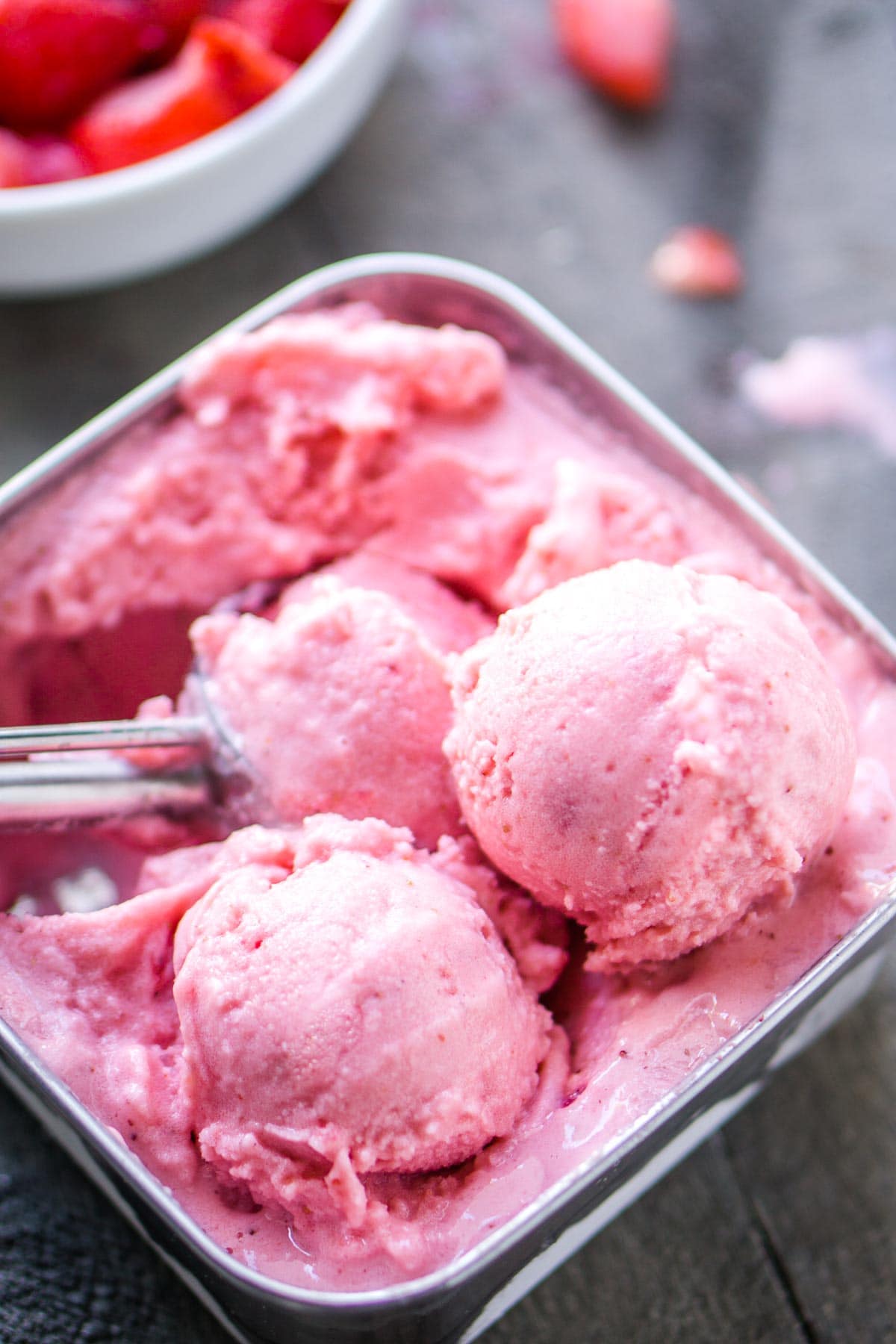 Three scoops of pink strawberry frozen yogurt in a metal container with a scooper, with a bowl of strawberries in the background—perfect inspiration if you’re learning how to make frozen yogurt at home.