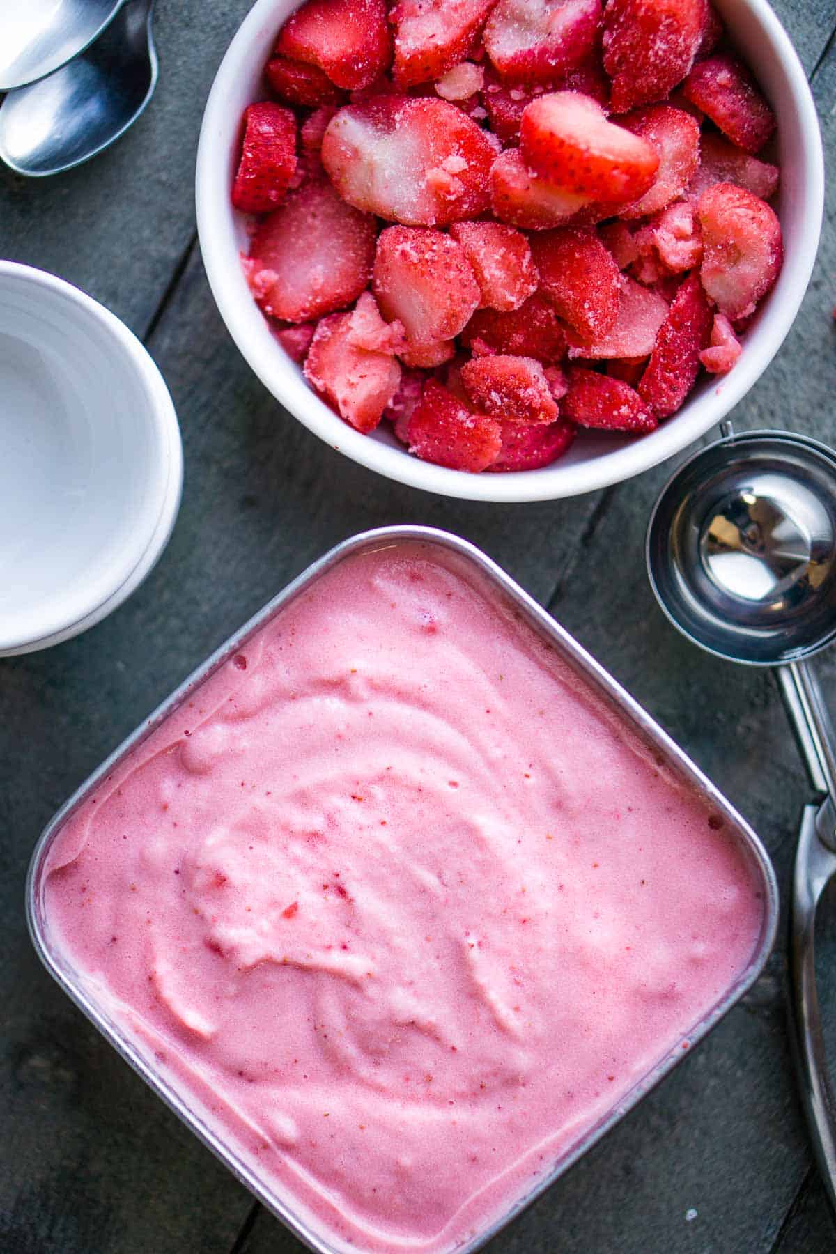 strawberry frozen yogurt in square storage container next to ice cream scoop and bowl of frozen strawberries
