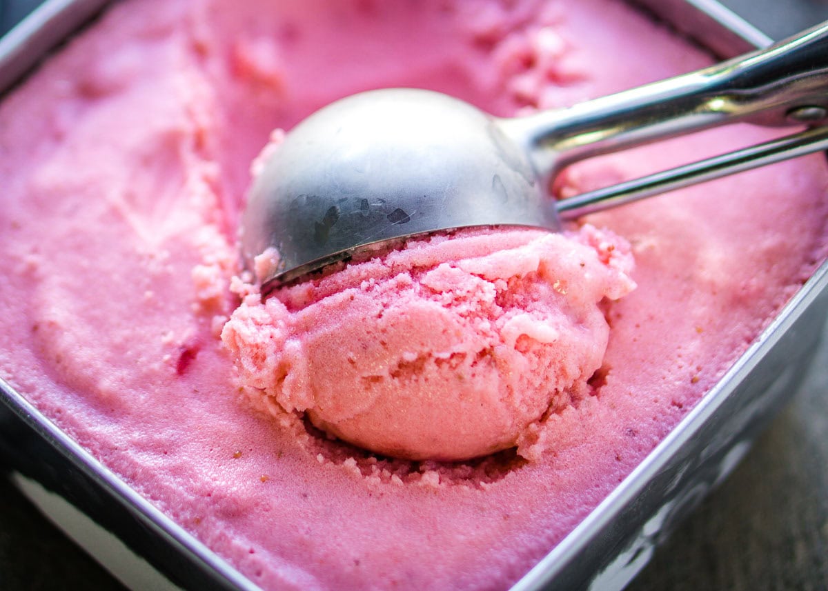 A metal ice cream scoop is lifting a scoop of pink strawberry frozen yogurt from a container.