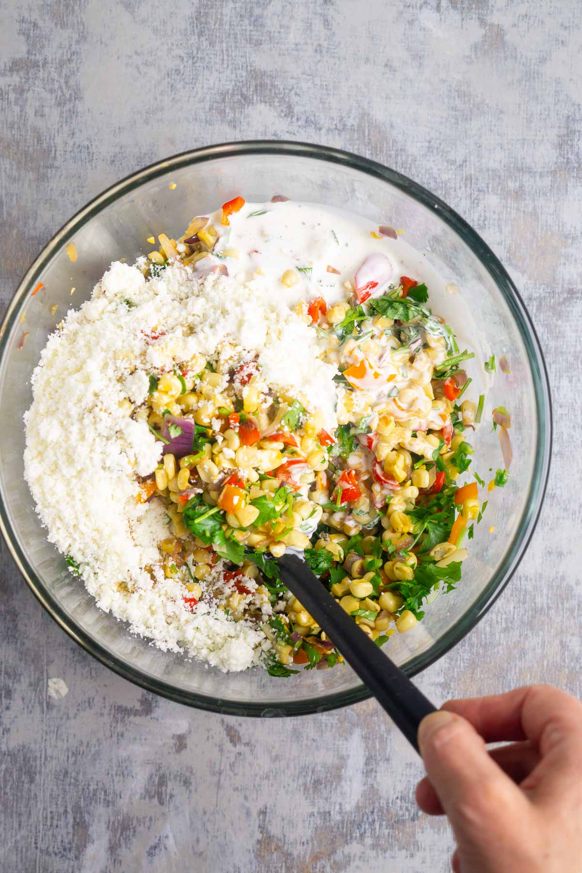 Person's hand uses black stirring utensil to mix ingredients for street corn salad in a glass mixing bowl.