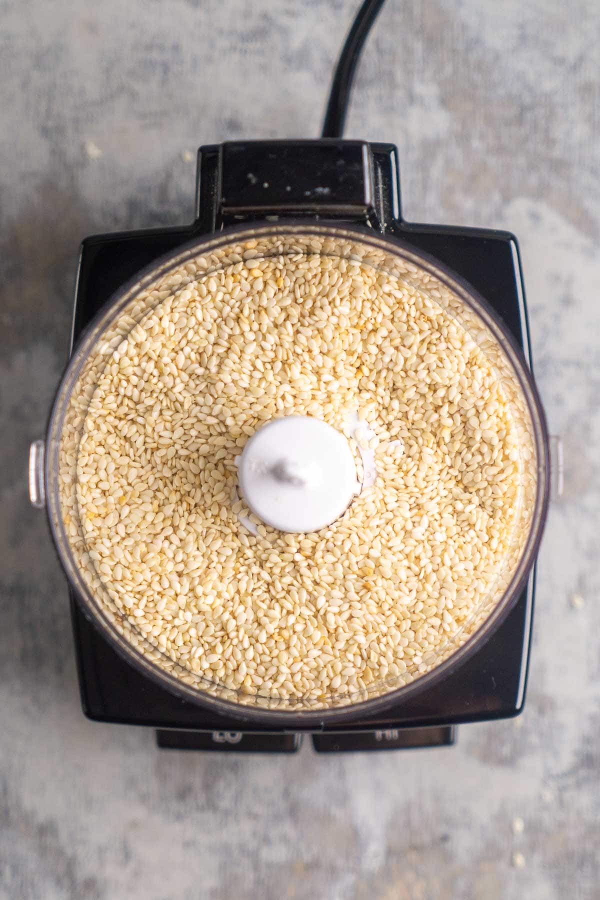 Top view of a food processor filled with white sesame seeds on a textured surface.