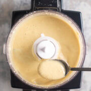 A food processor containing creamy tahini paste. A spoon is lifting some of the paste above the processor.