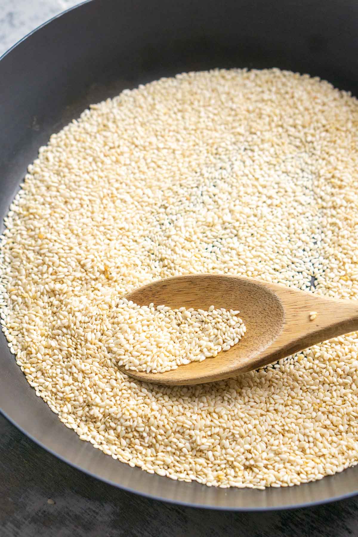 White sesame seeds in a black pan with a wooden spoon scooping them.