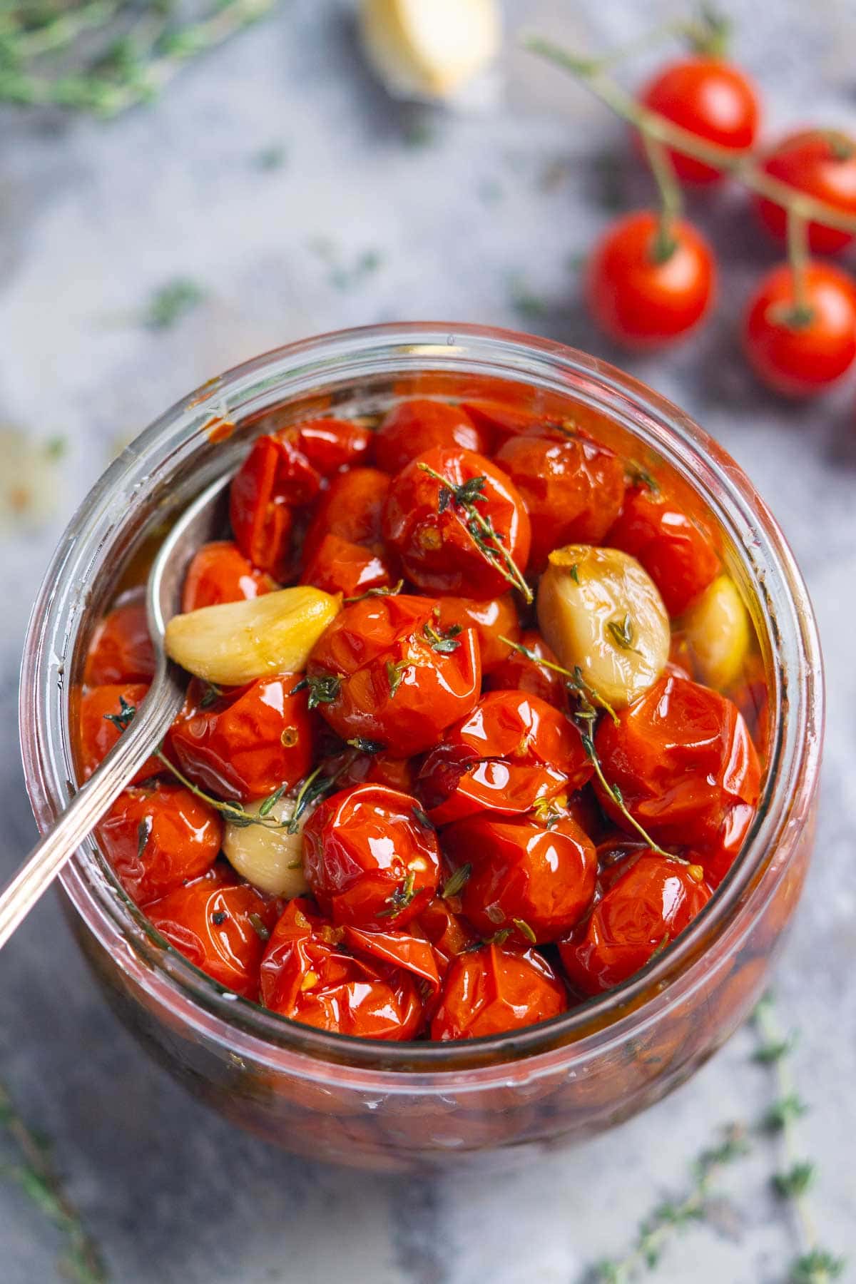 A glass jar filled with cherry tomato confit—roasted cherry tomatoes, whole garlic cloves, and herbs—with a spoon resting inside. Some cherry tomatoes and garlic are visible in the background.