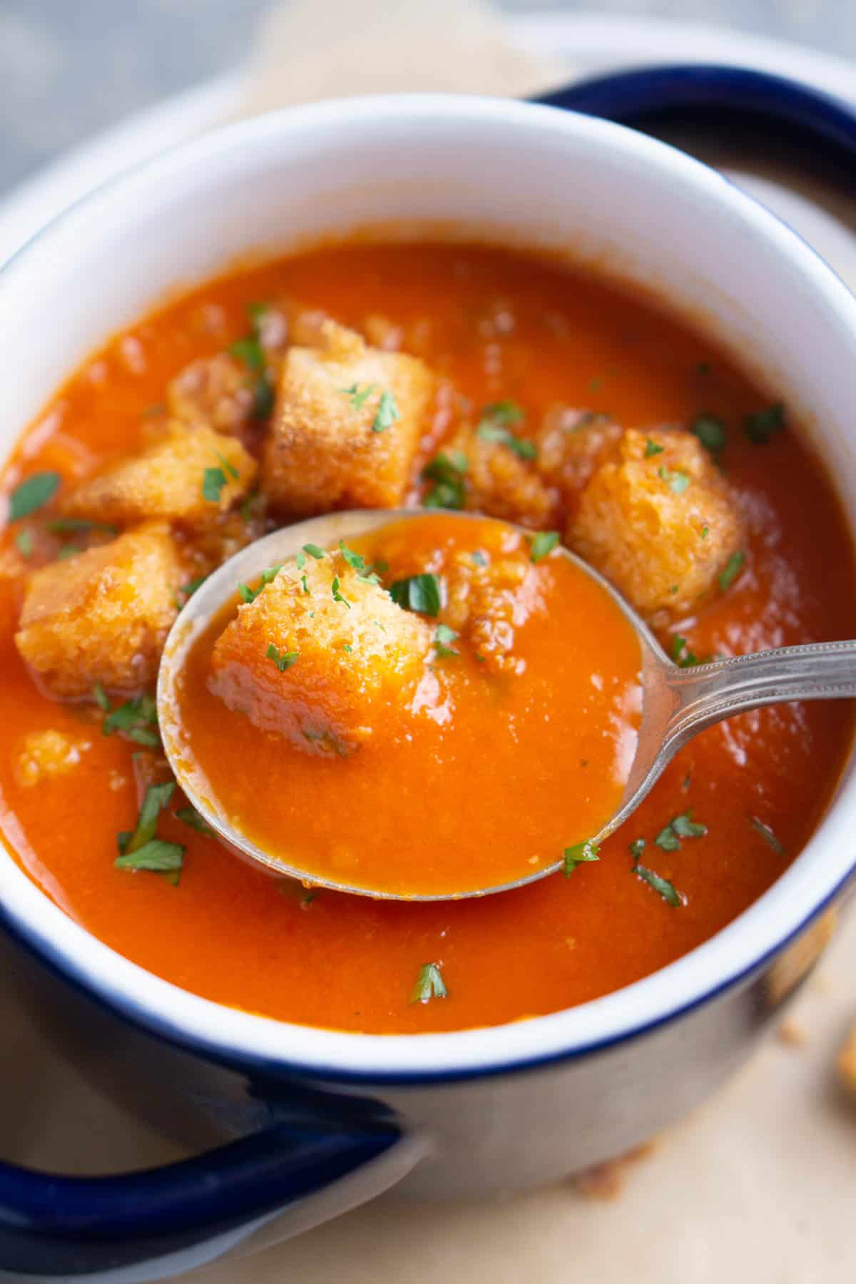 soup spoon lifts tomato soup from blue and white crock topped with croutons and parsley