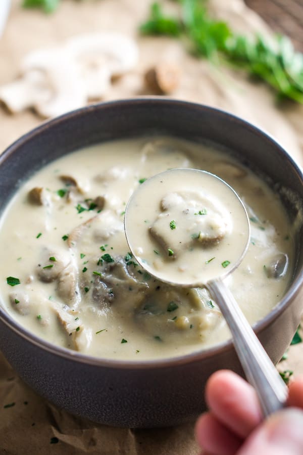 Soup spoon picks up vegan cream of mushroom soup from brown ceramic bowl on crinkled brown paper with mushroom, parsley, and nutmeg garnish