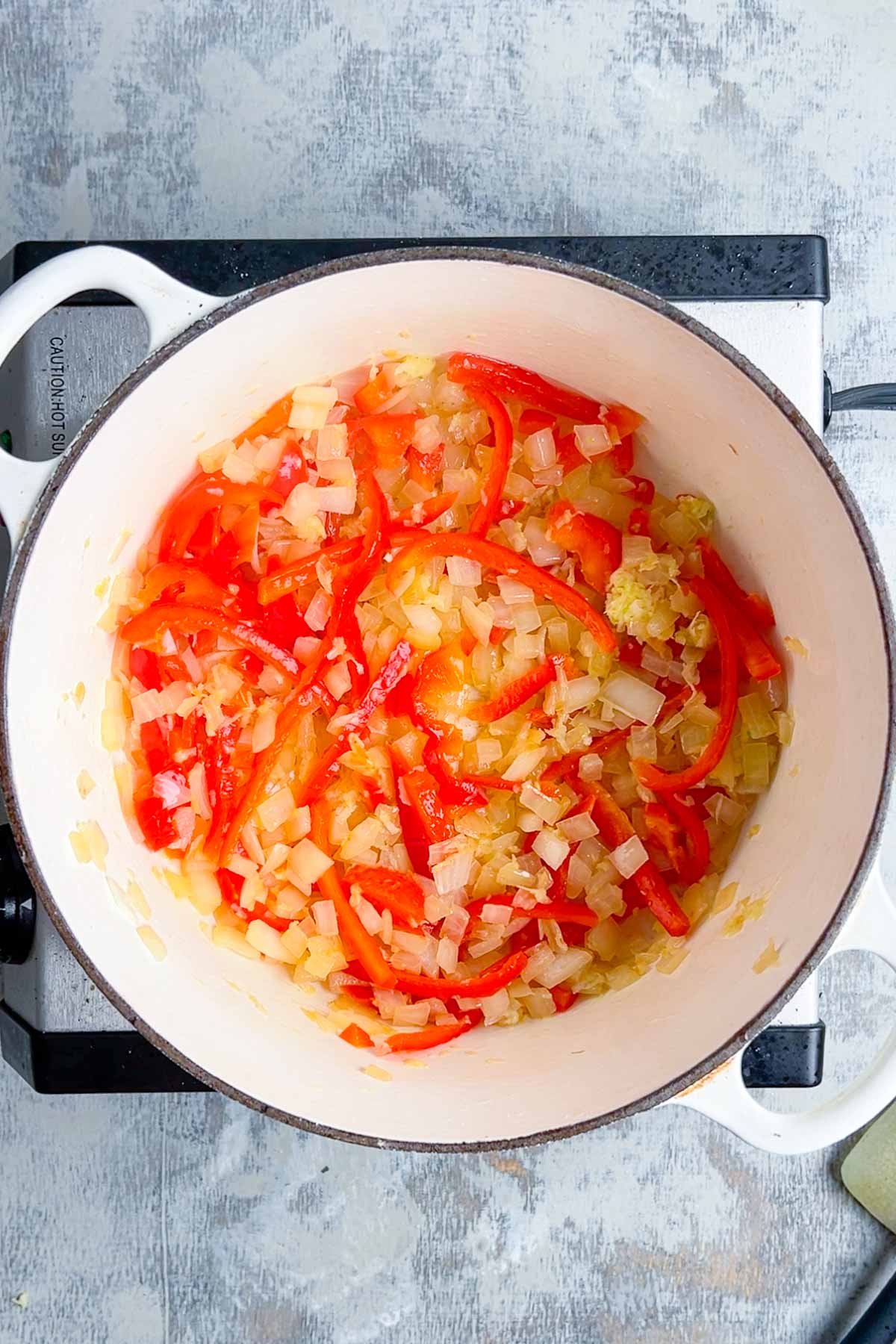 Chopped onions and sliced red peppers cooking in a white pot on a stovetop.