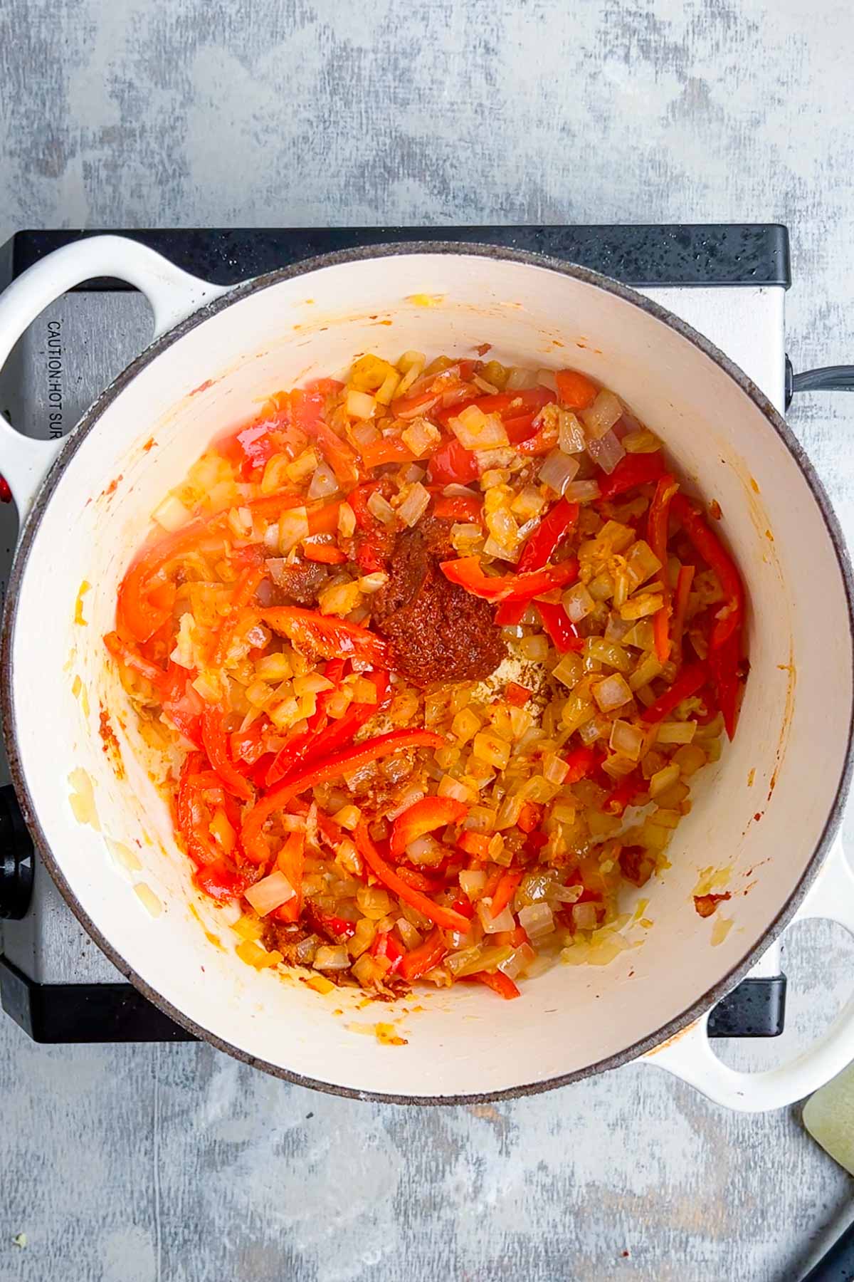 A white pot on a stovetop contains chopped onions and red bell peppers. Seasonings are sprinkled on top. The vegetables appear to be sautéing.