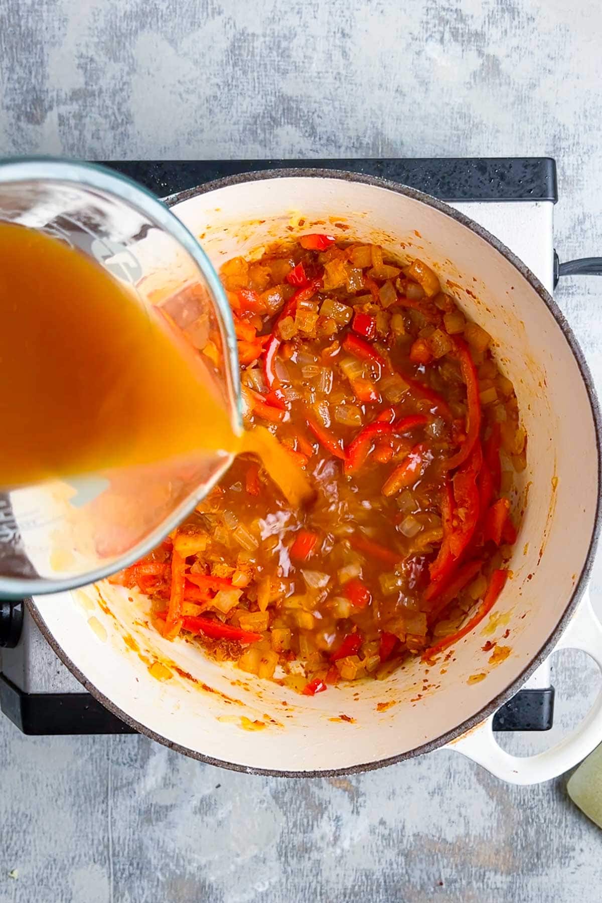 Pouring broth into a pot of sautéed onions and red peppers on a stove.