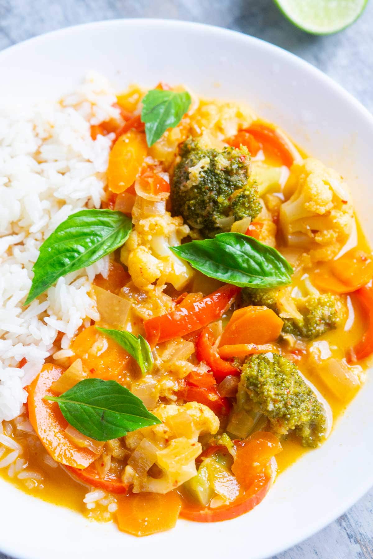 A plate of vegetable curry with broccoli, carrots, and peppers, garnished with basil leaves, served alongside steamed white rice.