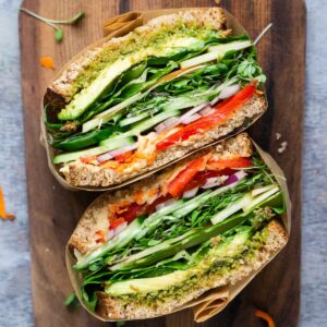 A parchment-wrapped sandwich filled with avocado, lettuce, cucumber, red pepper, onion, and sprouts is cut in half and placed on a wooden board.