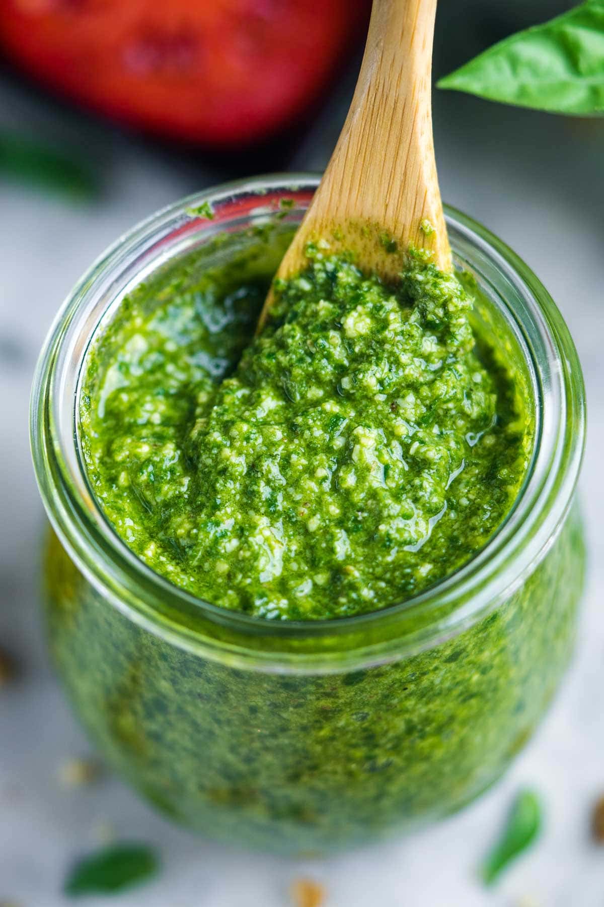 A glass jar filled with basil pesto sauce, with a wooden spoon scooping some out; fresh basil leaves are visible nearby.