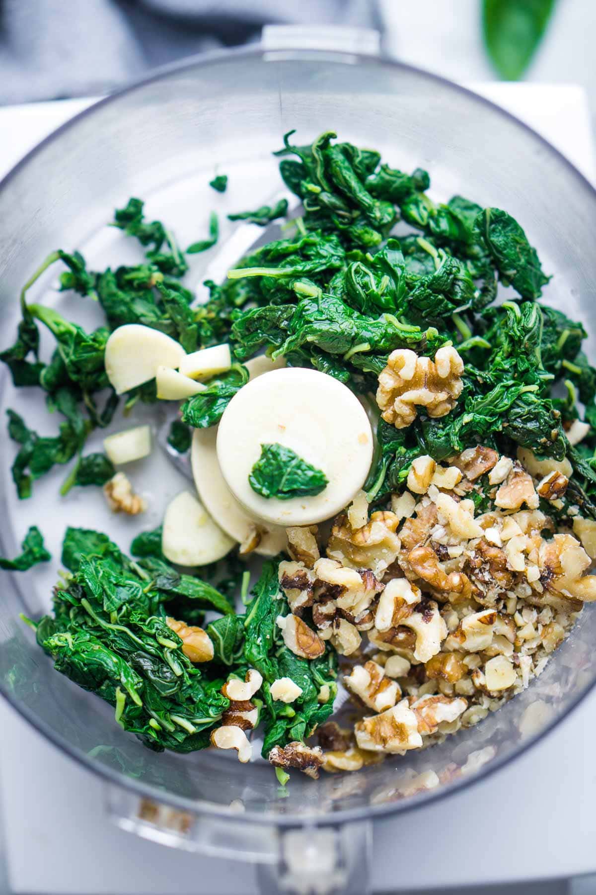 Blanched basil, walnuts, and garlic cloves inside a food processor bowl, ready to be blended for basil walnut pesto.