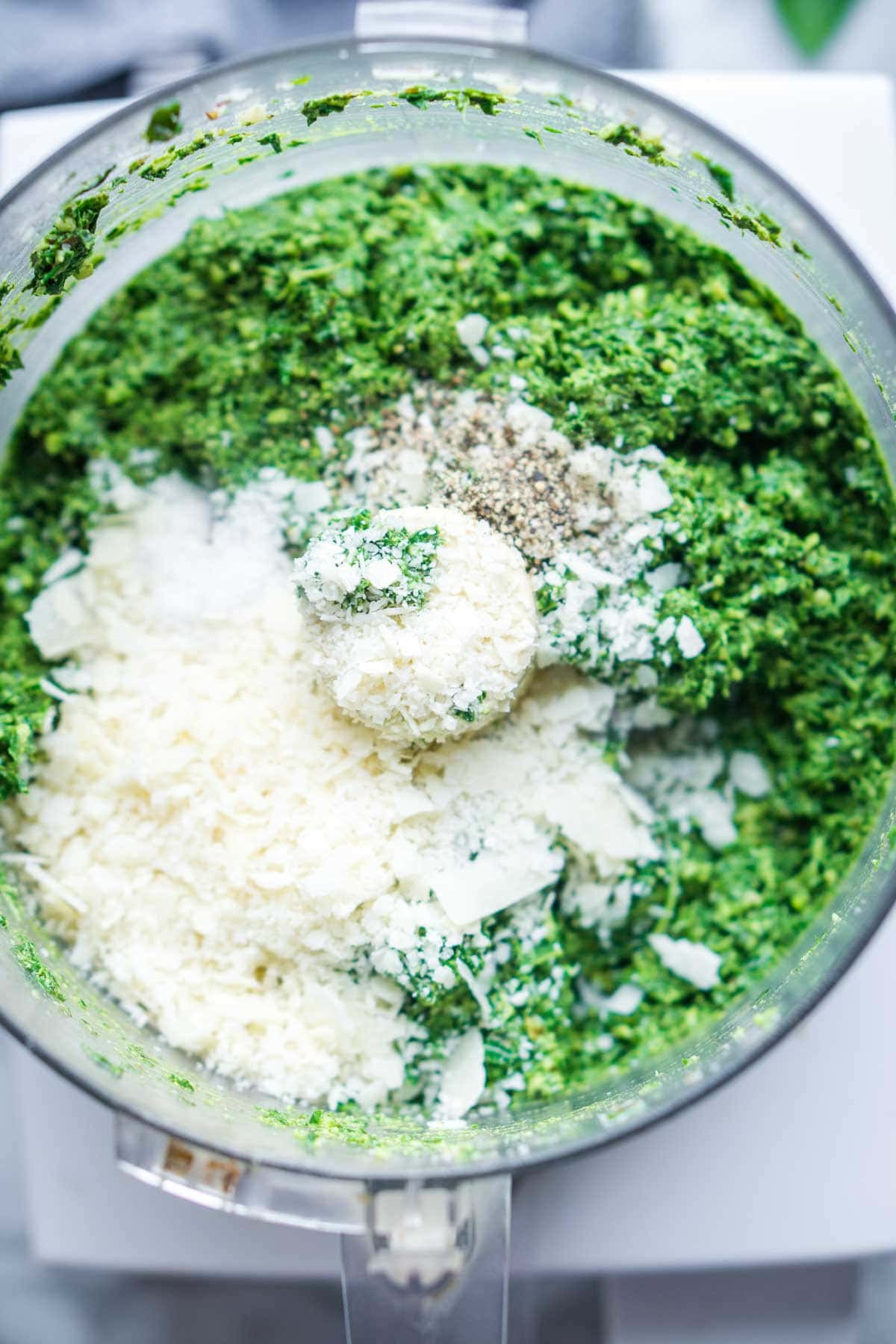 Grated Parmesan cheese on top of chopped basil and walnuts in a food processor bowl, ready to be blended into basil pesto.