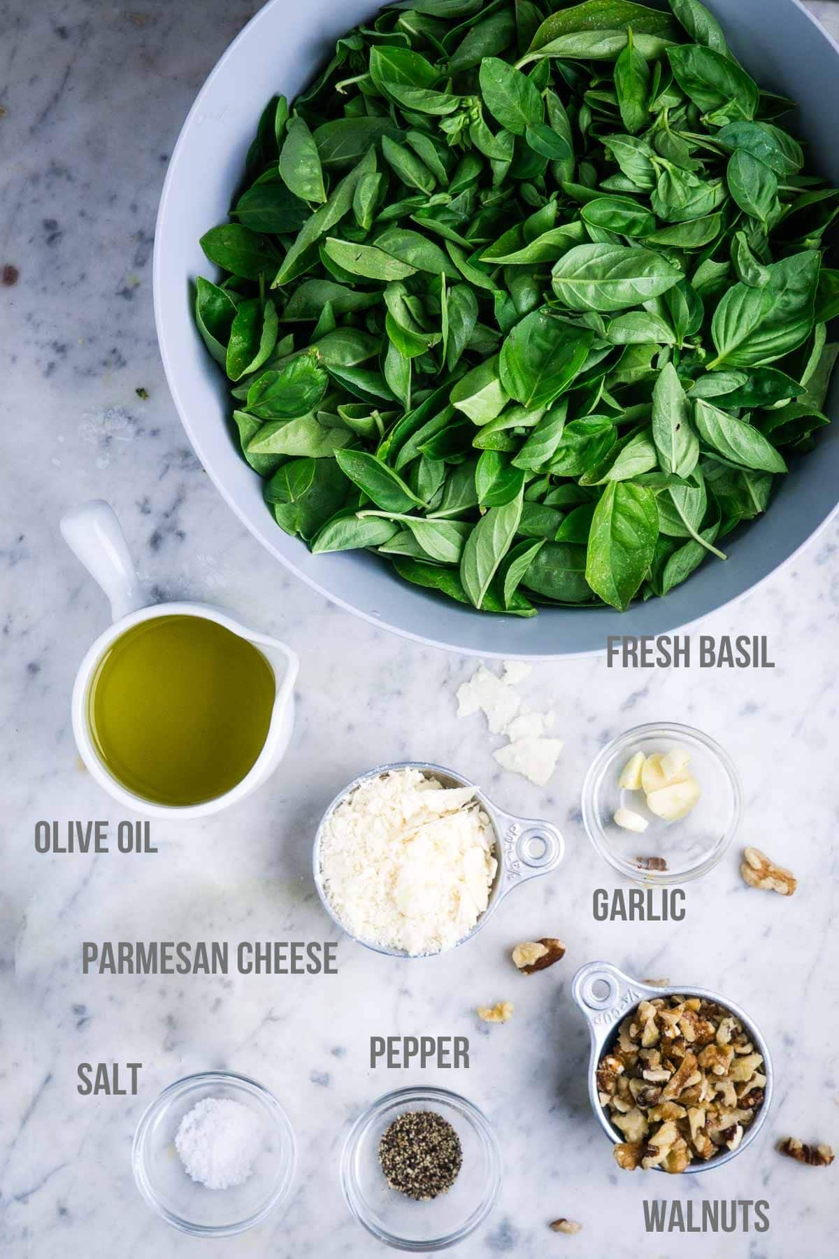 A bowl of fresh basil, a cup of olive oil, grated Parmesan cheese, salt, pepper, garlic cloves, and chopped walnuts arranged on a marble countertop in preparation for basil walnut pesto.