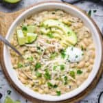white chicken chili topped with sliced avocado, shredded jack cheese, sour cream, and lime wedges in white bowl on cutting board trivet
