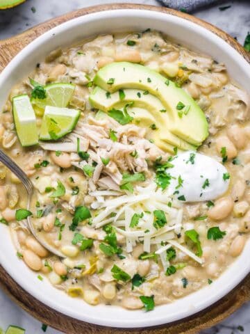white chicken chili topped with sliced avocado, shredded jack cheese, sour cream, and lime wedges in white bowl on cutting board trivet