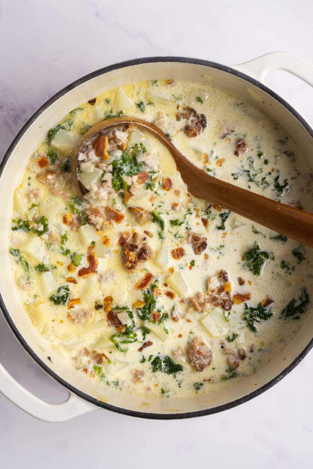 Large white pot filled with creamy zuppa toscana soup containing sausage, kale, potatoes, and bacon bits. A wooden spoon rests inside the pot on a light surface.