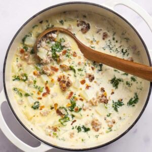 A pot of creamy zuppa toscana soup with sausage, potatoes, kale, and bacon, stirred with a wooden spoon.
