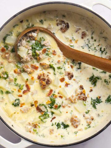 A pot of creamy zuppa toscana soup with sausage, potatoes, kale, and bacon, stirred with a wooden spoon.