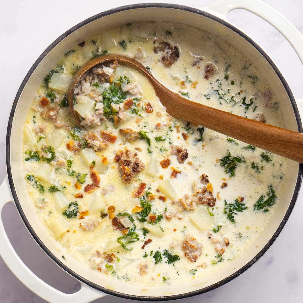 A pot of creamy zuppa toscana soup with sausage, potatoes, kale, and bacon, stirred with a wooden spoon.