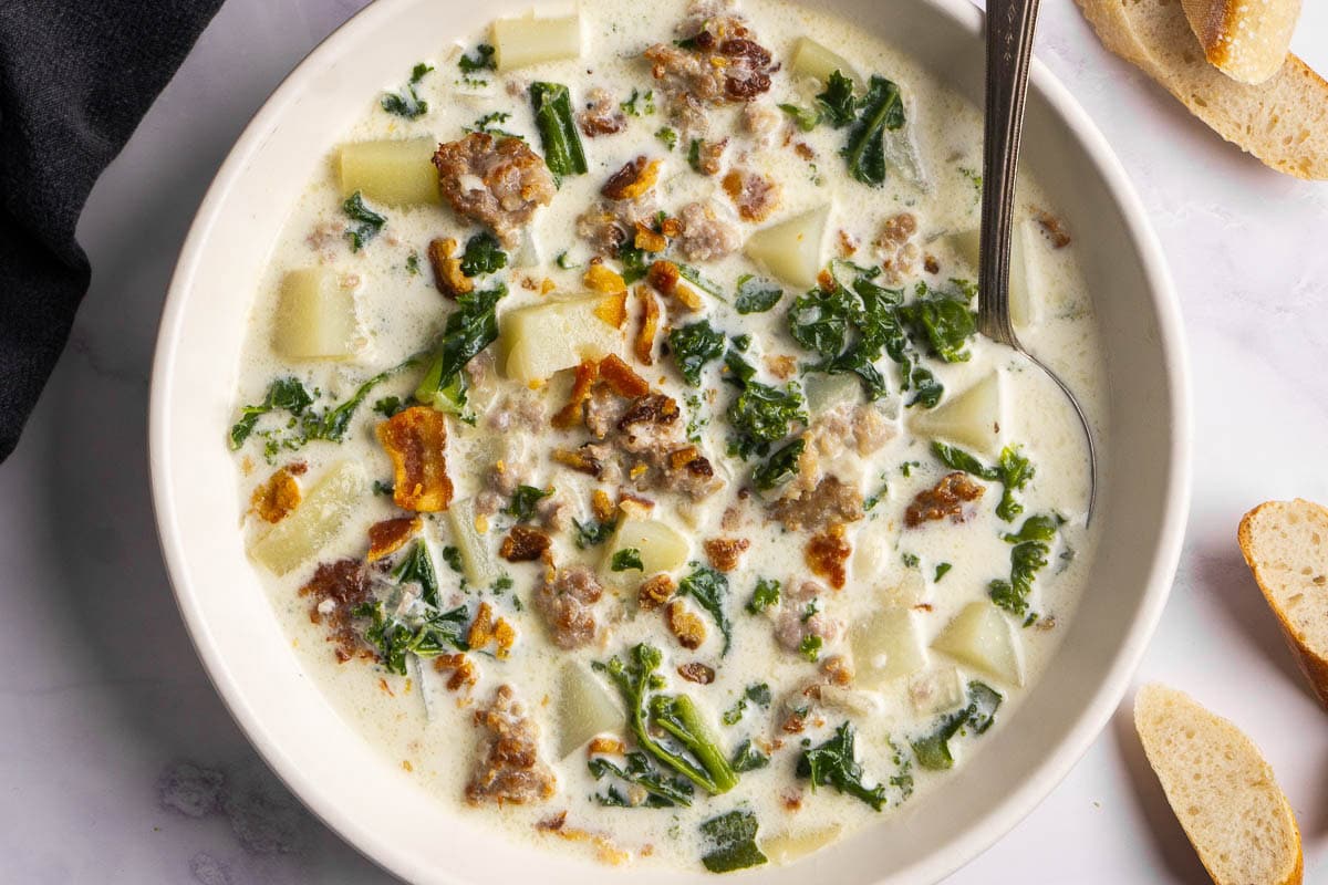 A bowl of creamy zuppa toscana soup with sausage, potatoes, kale, and bacon pieces, with a spoon in the bowl and slices of bread on the side.