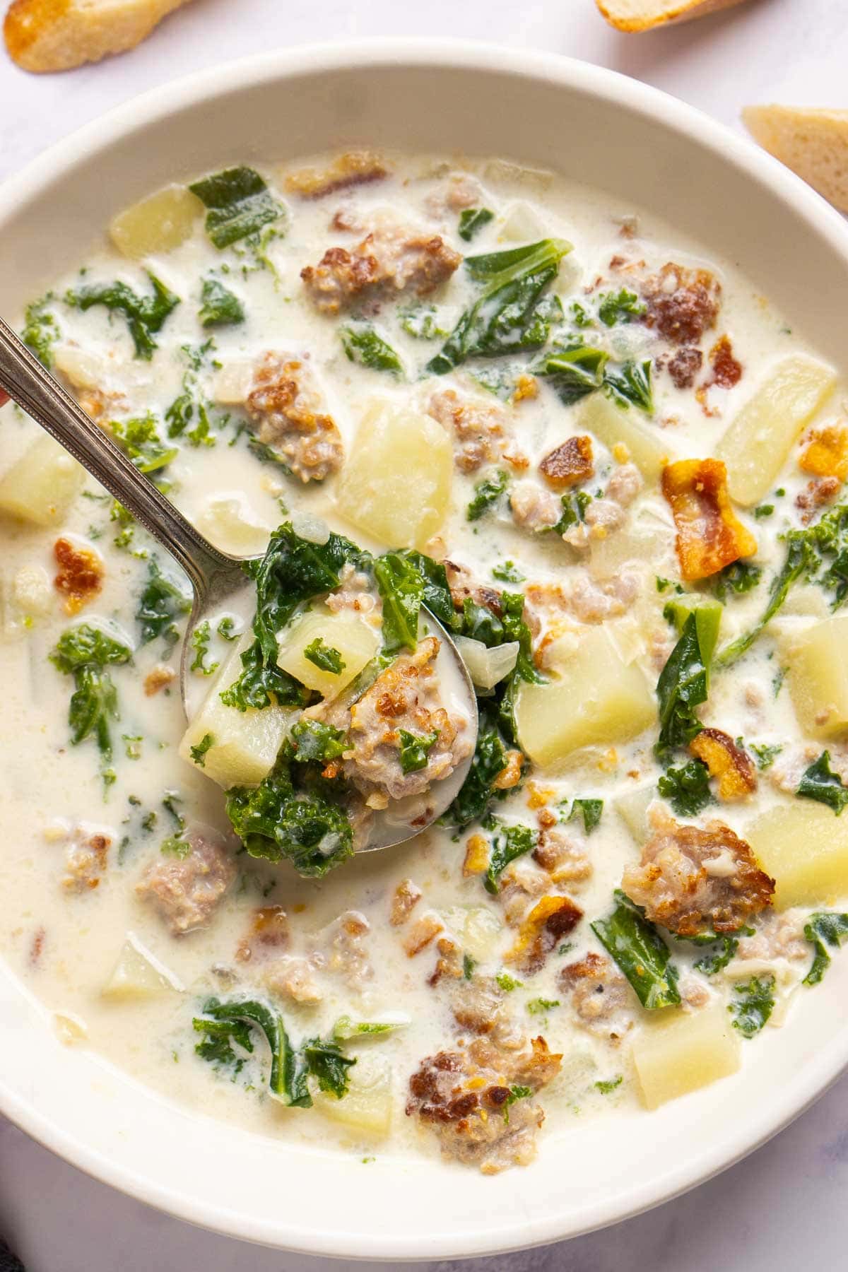 A bowl of creamy zuppa toscana soup with chunks of sausage, potatoes, and kale, with a spoon lifting a portion from the bowl.
