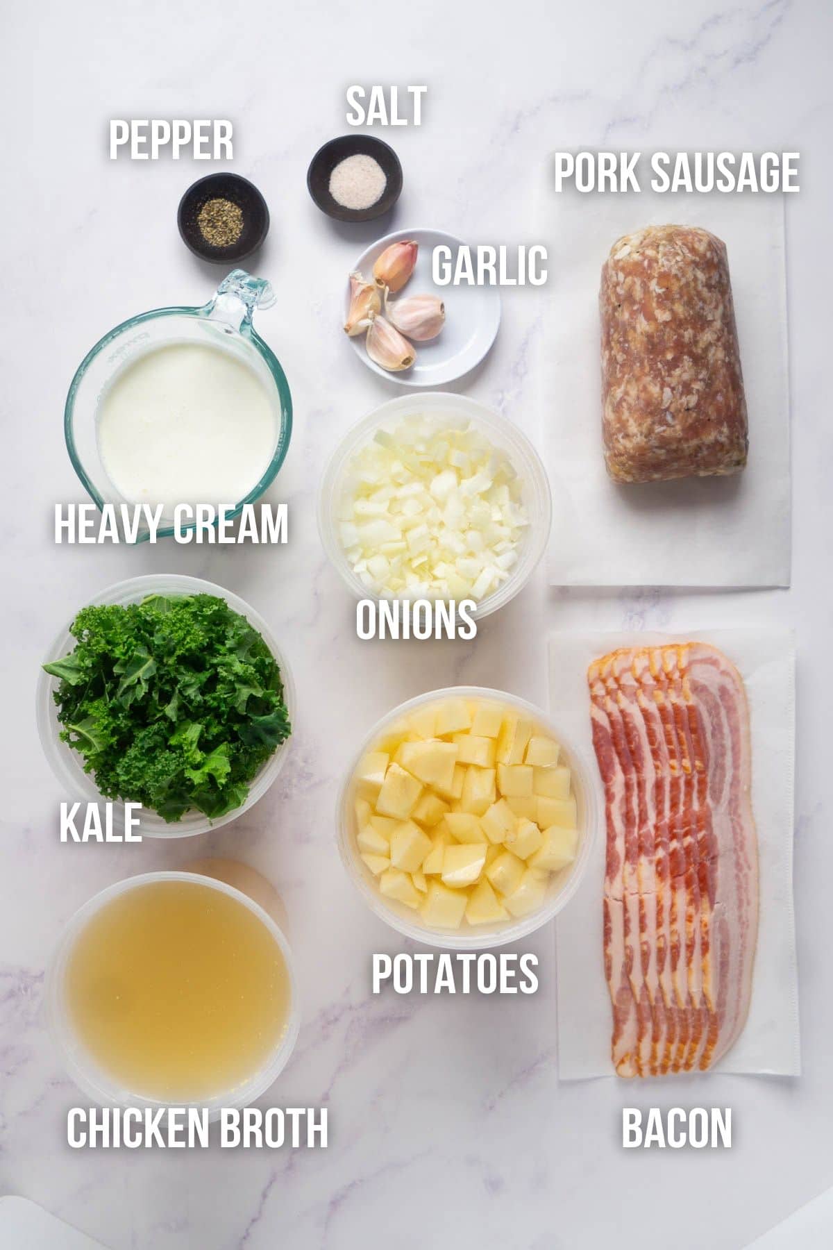 Ingredients laid out on a white surface: pork sausage, bacon, potatoes, onions, kale, chicken broth, heavy cream, garlic, salt, and pepper.